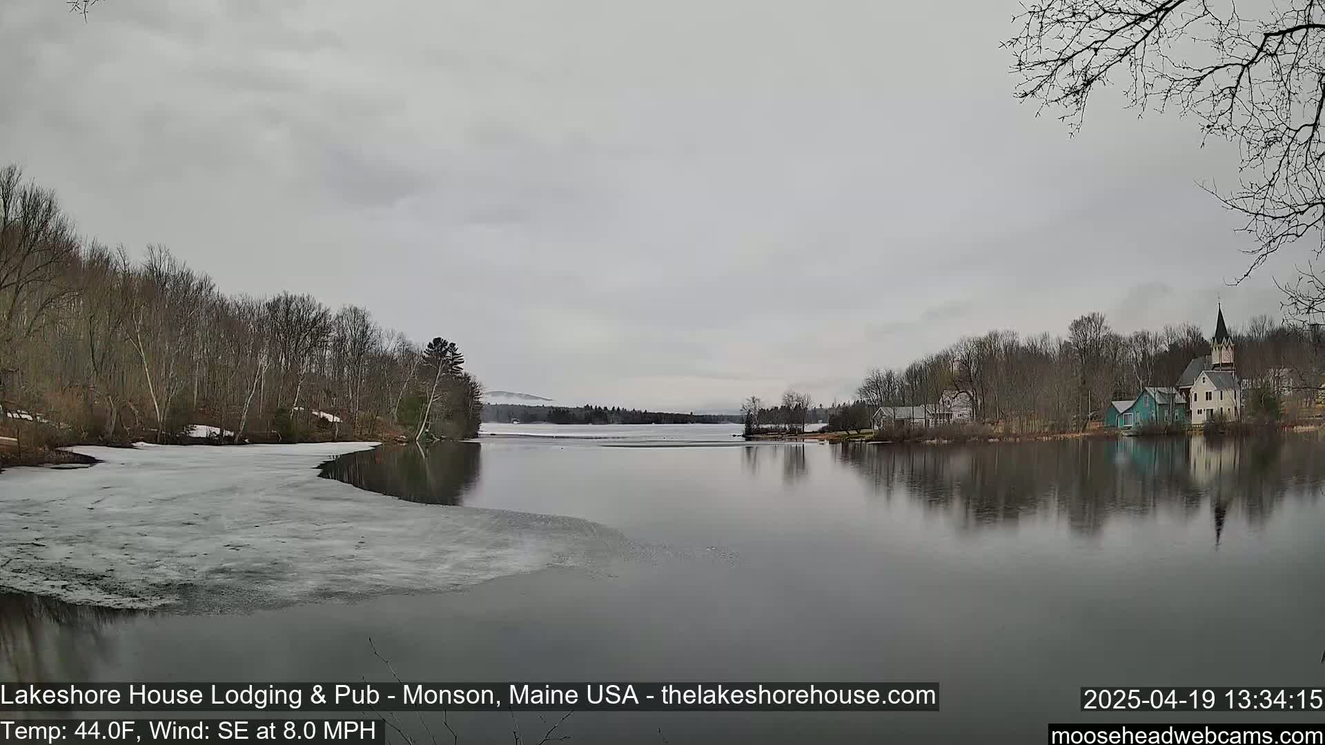 A calm lake with some remaining ice, partially surrounded by bare trees and a small village under an overcast sky.