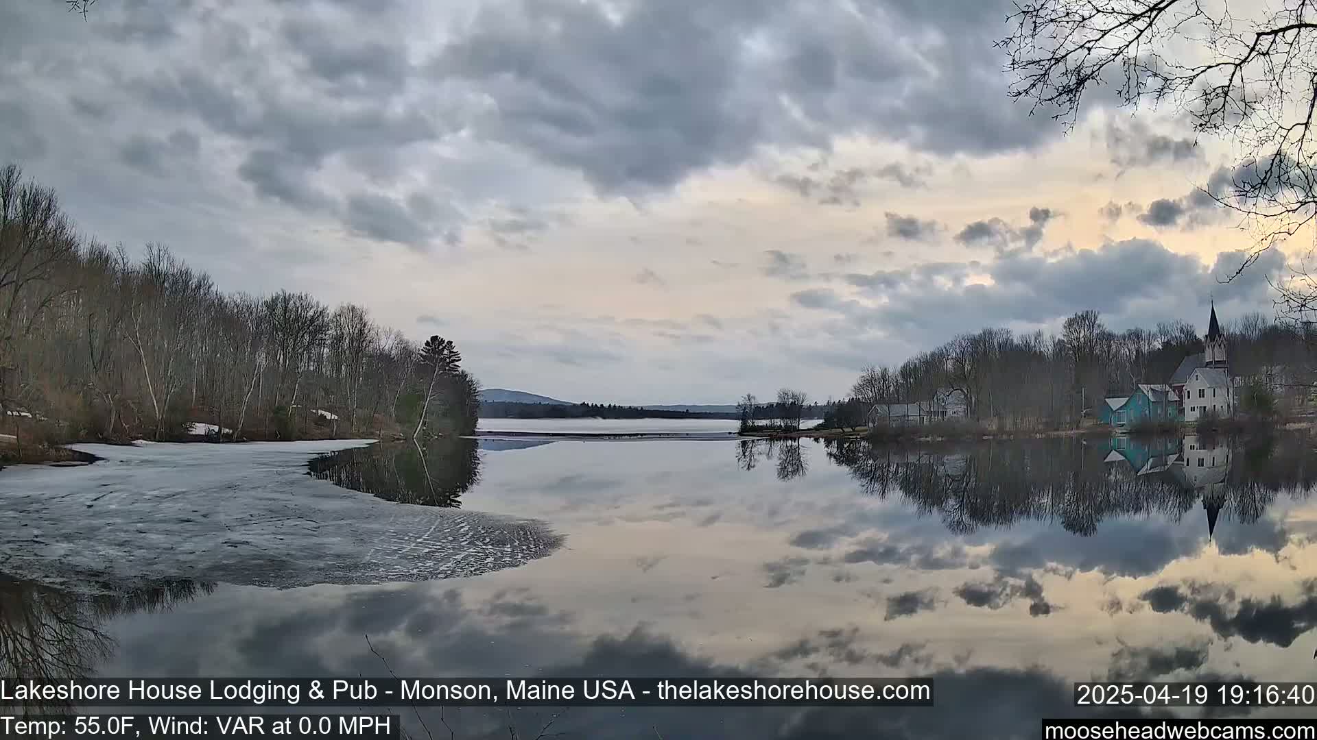 A calm lake with partially frozen edges reflects a cloudy sky and a small town nestled among bare trees.