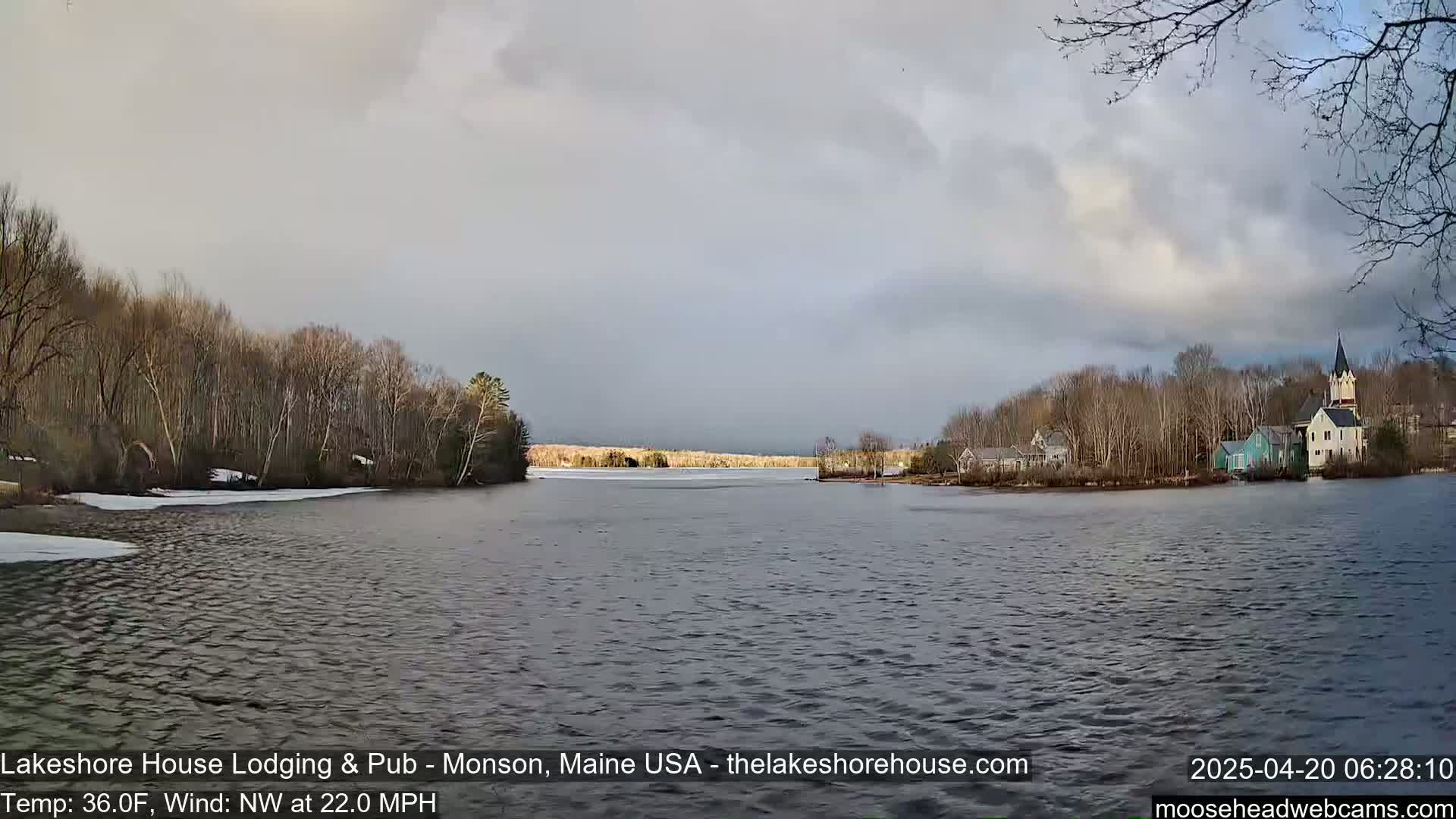 A mostly cloudy sky hangs over a calm lake with a partially frozen shoreline, bare trees, and a village of buildings on the far side.