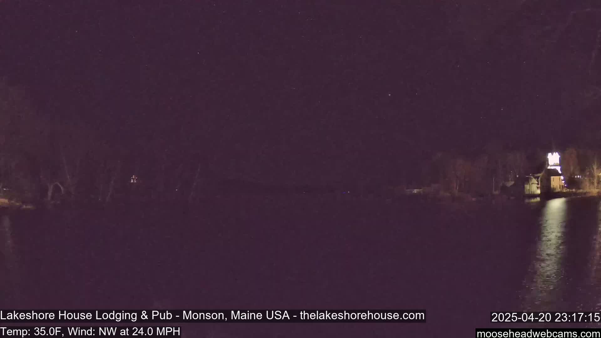 A nighttime view of a calm body of water with a building illuminated on the opposite shore under a starry sky, with a northwest wind at 24 mph and a temperature of 35 degrees Fahrenheit.