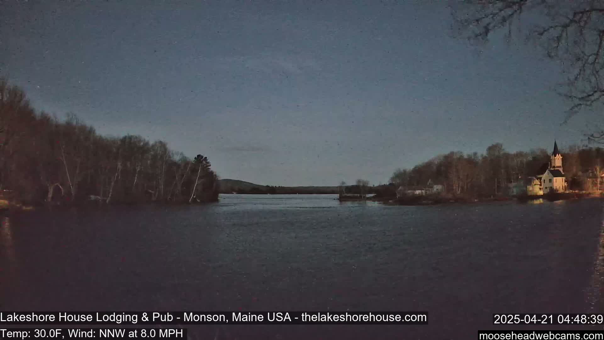 A dark, calm lake is seen at twilight, with trees and buildings on the far shore under a clear, dark sky.
