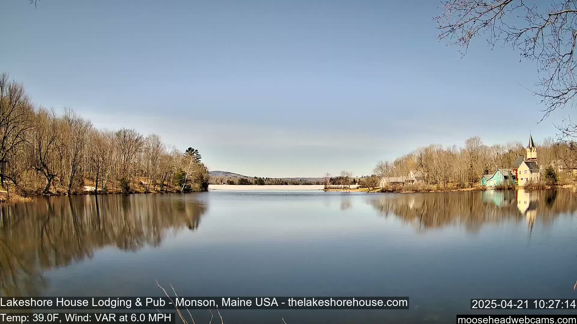 A calm lake reflects leafless trees and a few buildings on a sunny day with a light breeze.
