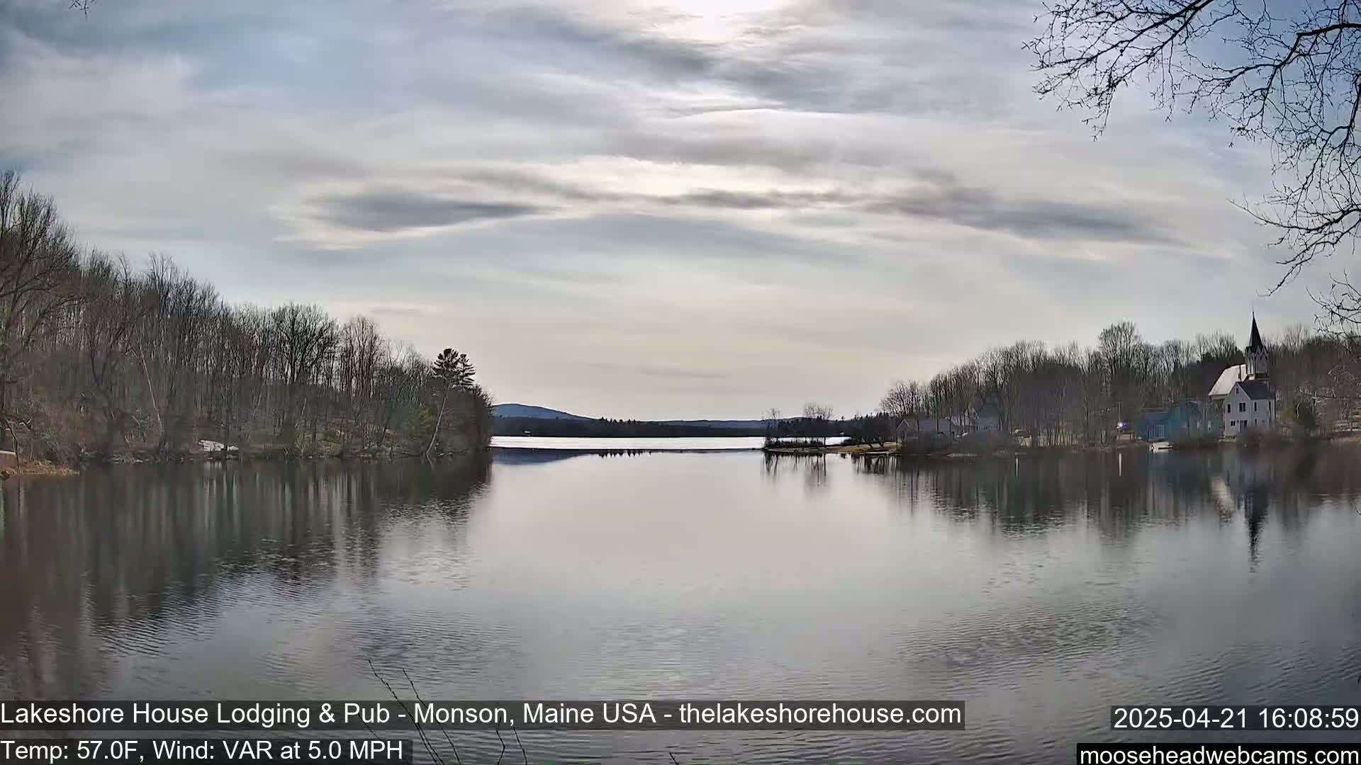 A calm lake reflects a cloudy sky and bare trees, with buildings and a church steeple visible on the far shore.