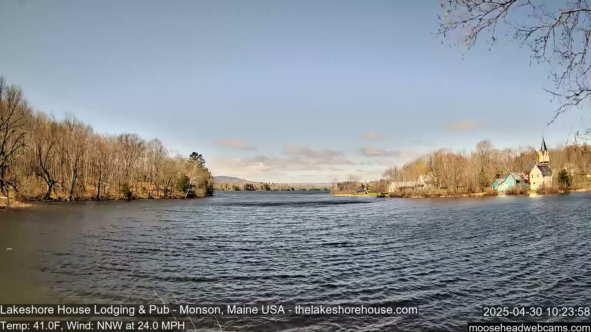 A mostly calm lake is seen under a clear, bright sky, with bare trees lining the shores and a few buildings visible in the distance on the right.
