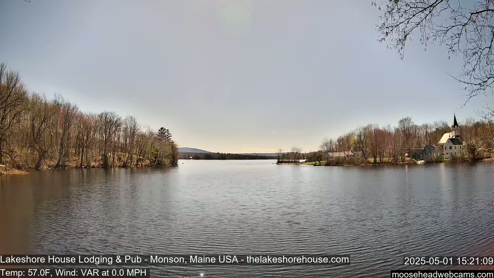 A calm lake is surrounded by bare trees and a few buildings, under a mostly clear sky.