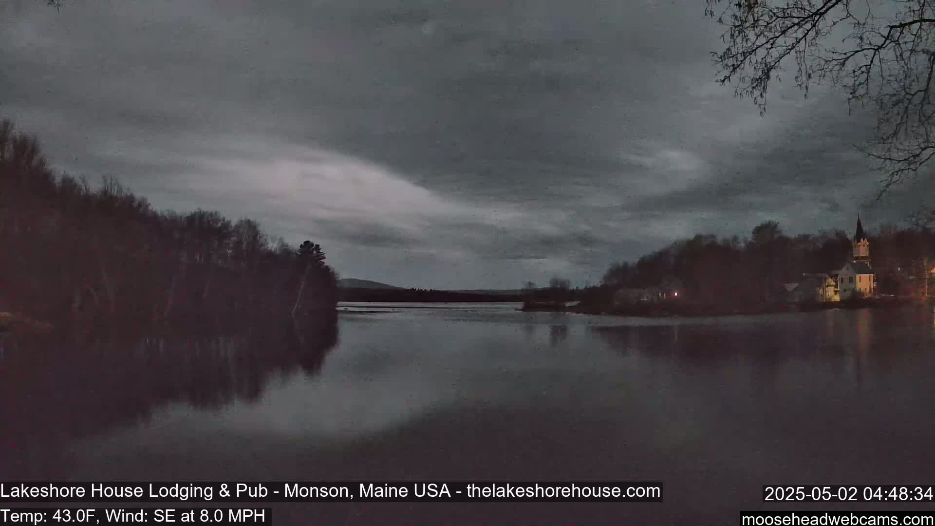 A calm, dark lake is reflected under an overcast sky, with trees and a building visible on the far shore.