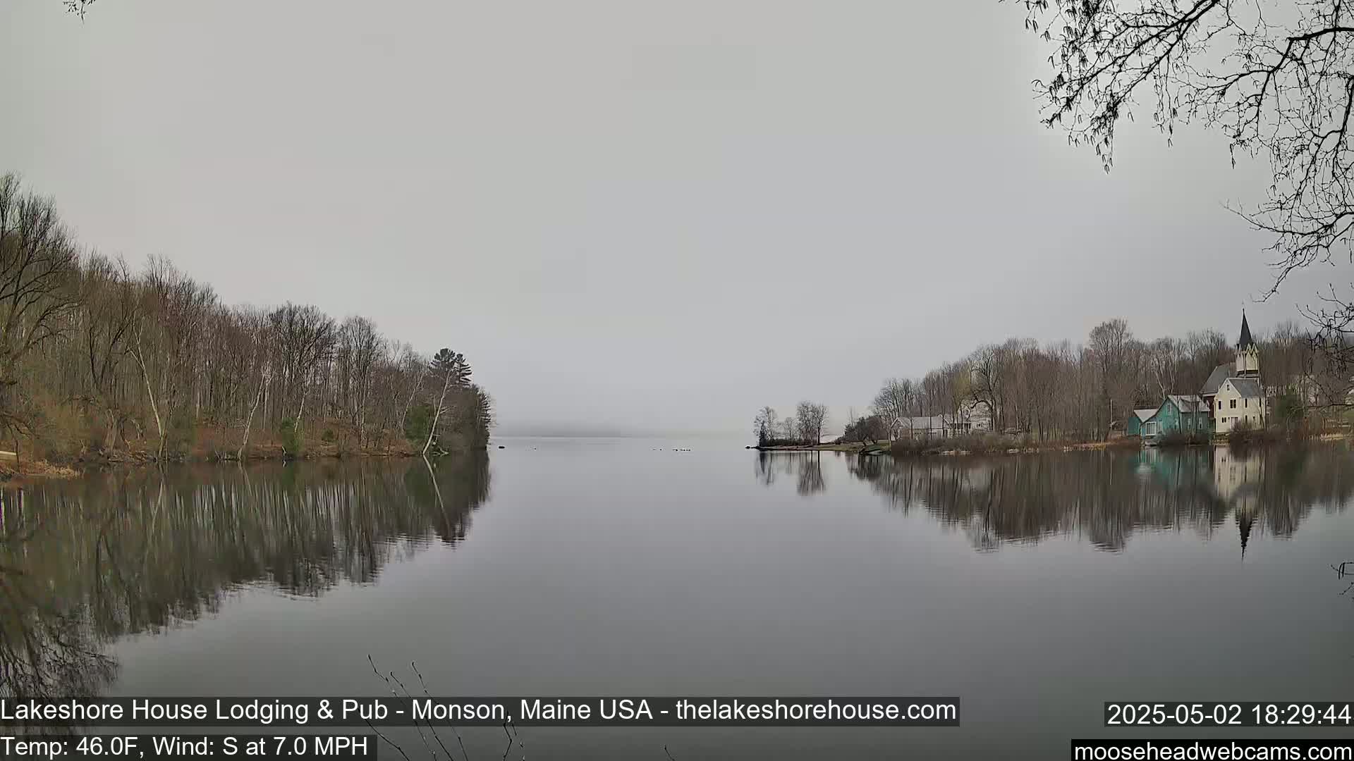 A calm lake reflects a mostly bare, tree-lined shoreline with a few buildings, under an overcast sky.