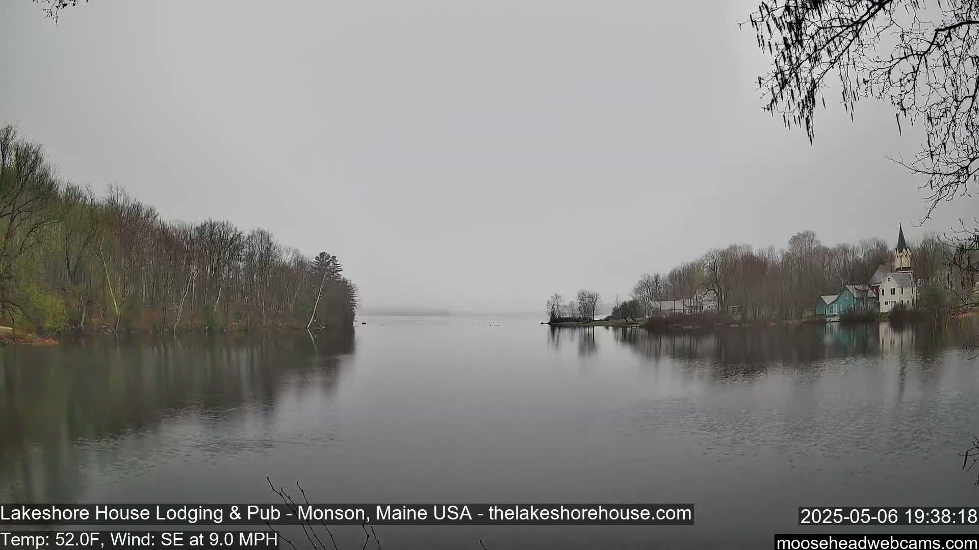 A calm lake, under an overcast sky, is surrounded by bare trees and a few buildings on the far shore.