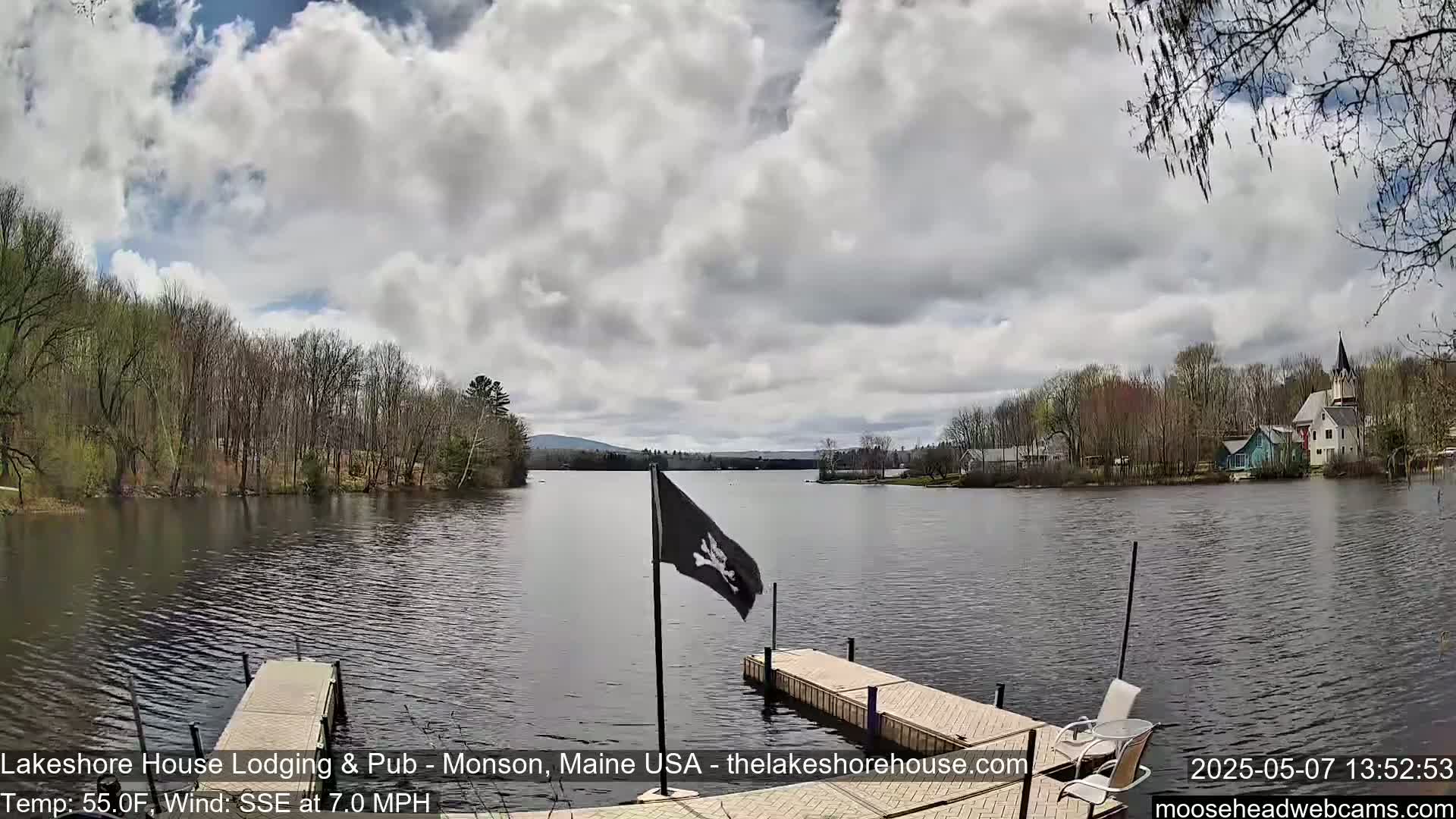 A calm lake is seen from a dock, under a partly cloudy sky, with trees and buildings lining the shore.