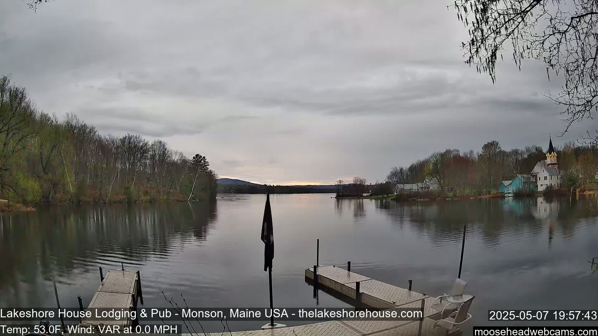 A calm lake is seen under an overcast sky, with trees lining the shore and buildings visible in the distance.