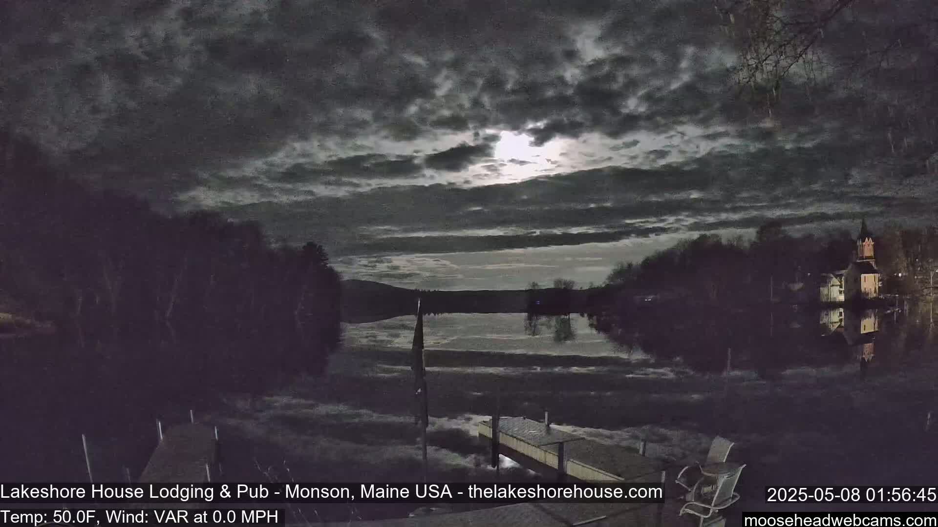 A nighttime view of a calm lake under a mostly cloudy sky, illuminated by a bright moon, shows dark trees lining the shore with buildings faintly visible in the distance.