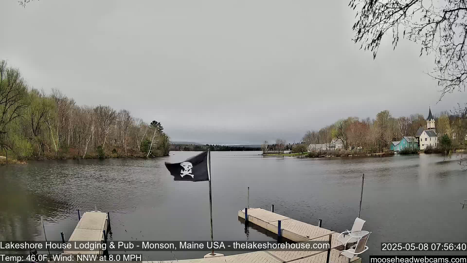 A calm lake with a small town on the opposite shore is viewed from a dock with a pirate flag, under an overcast sky.