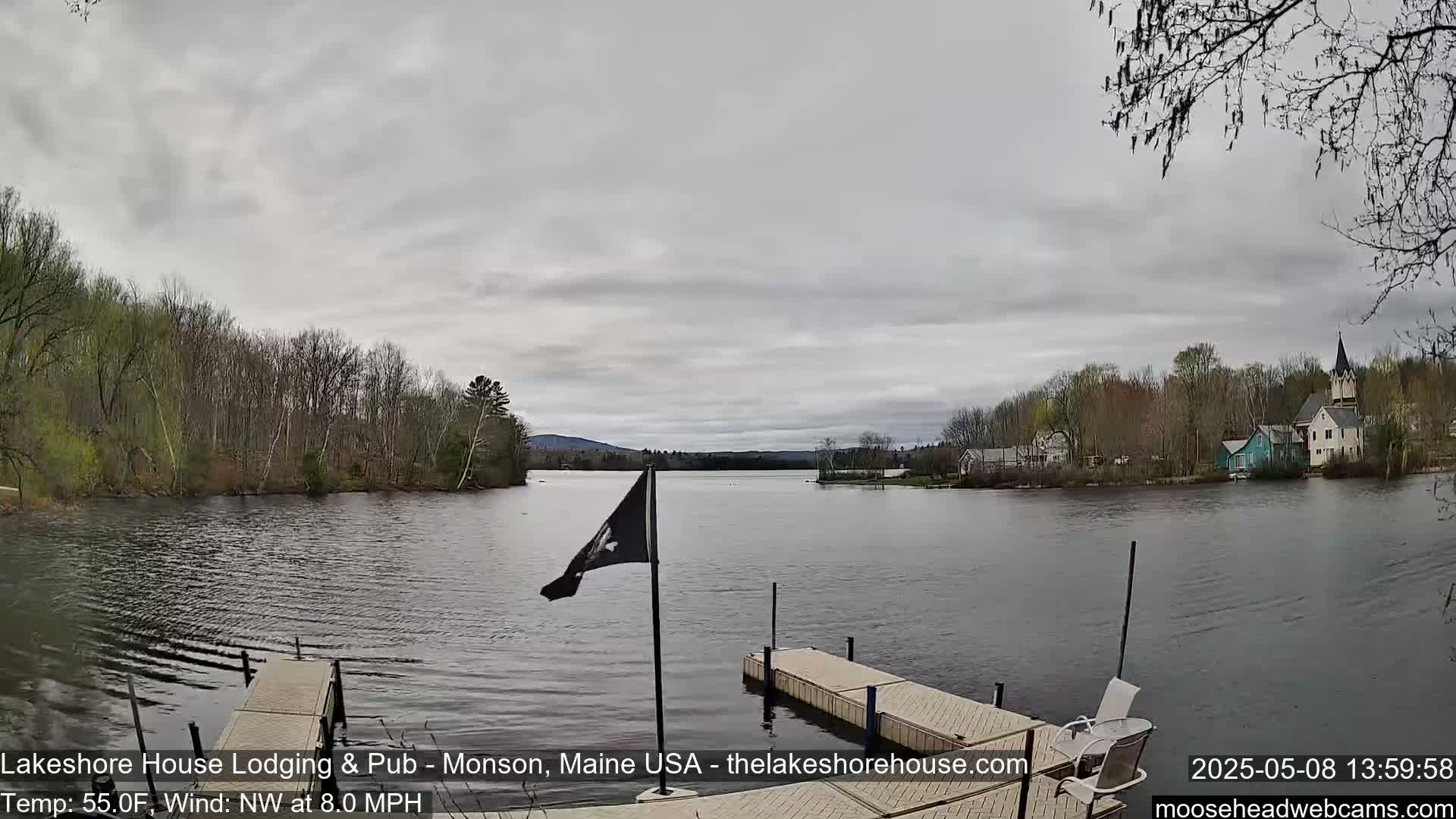 A placid lake under a cloudy sky is viewed from a wooden dock, with a small town and forested hills visible in the distance.