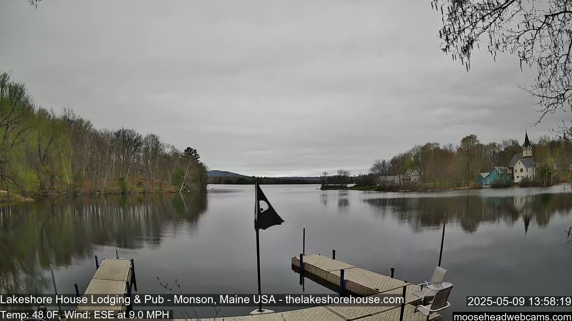 A calm lake reflecting a cloudy sky and spring foliage, with a small dock and distant buildings visible.