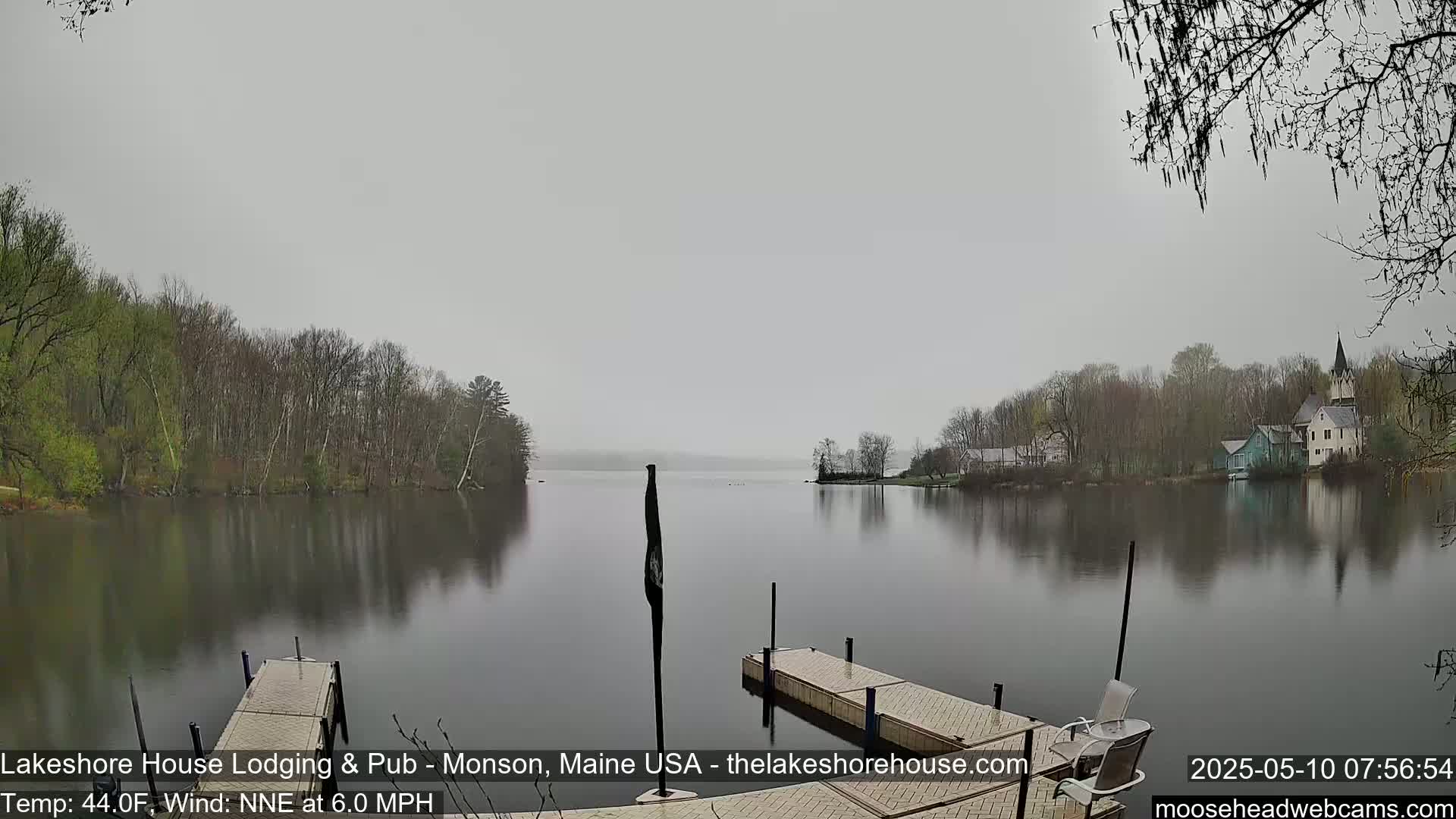 A calm lake is shrouded in a misty gray atmosphere, with trees lining the shore and a few buildings visible in the distance.