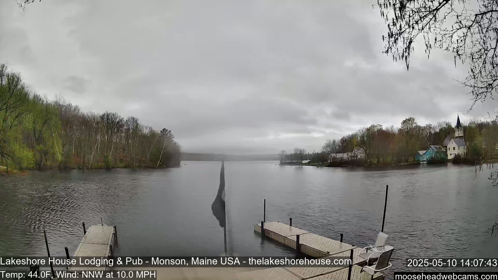 A calm lake is seen from a dock on an overcast day, with trees and buildings visible on the opposite shore.