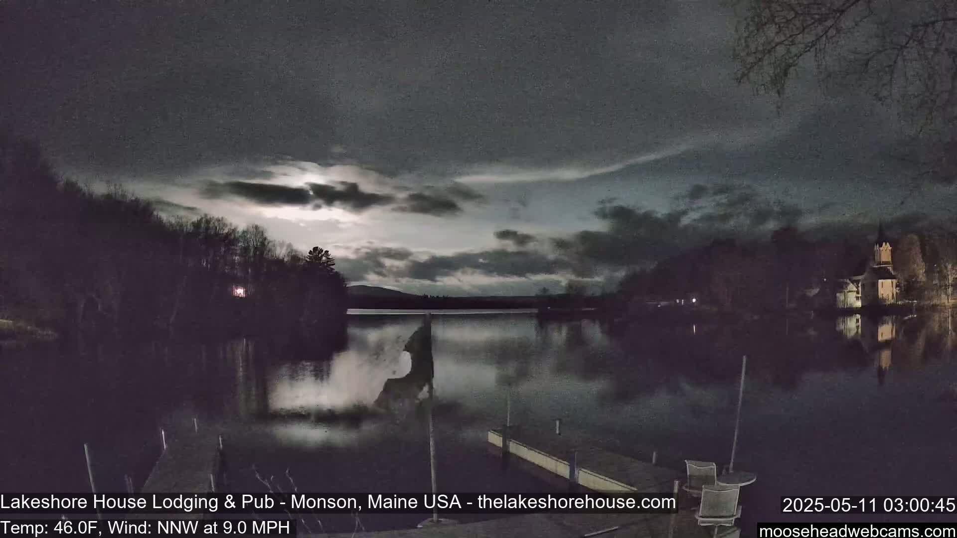 A moonlit lake is seen at night under a mostly cloudy sky, with trees and a building faintly visible across the water.