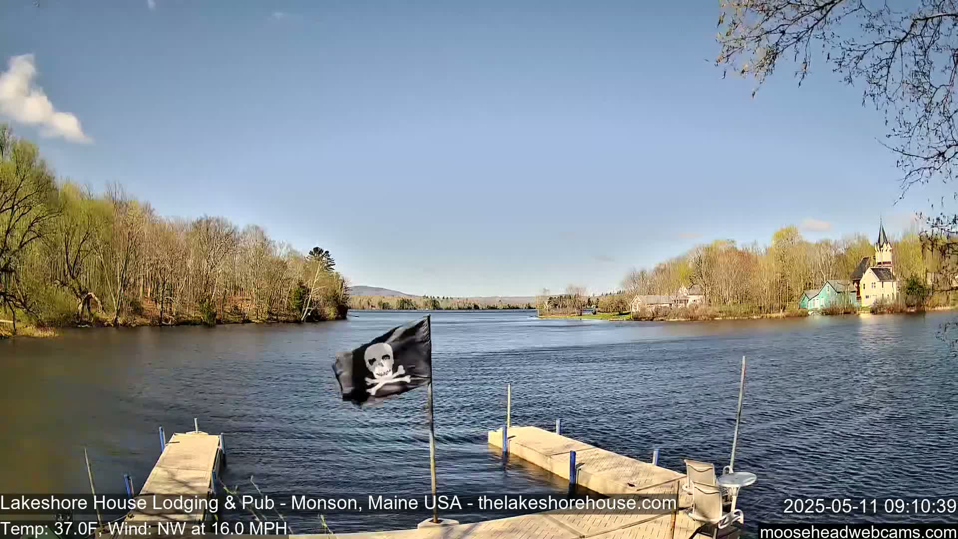 A calm lake with a pirate flag on a dock, surrounded by trees and buildings under a clear blue sky, with a northwest wind at 16 mph and a temperature of 37°F.