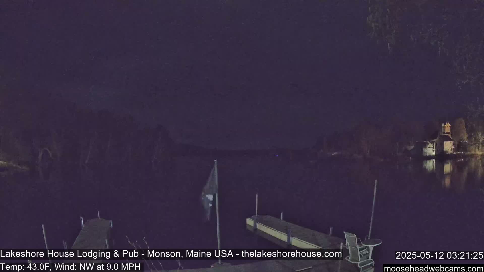 A nighttime view of a calm lake with a dock, a flagpole, and buildings faintly visible across the water under a dark sky, with a northwest wind at 9 mph and a temperature of 43°F.