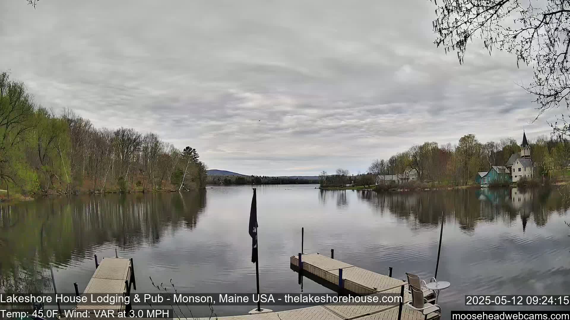 A calm lake reflects a cloudy sky and the spring foliage of trees lining its shores, with a few buildings visible across the water and a dock in the foreground.