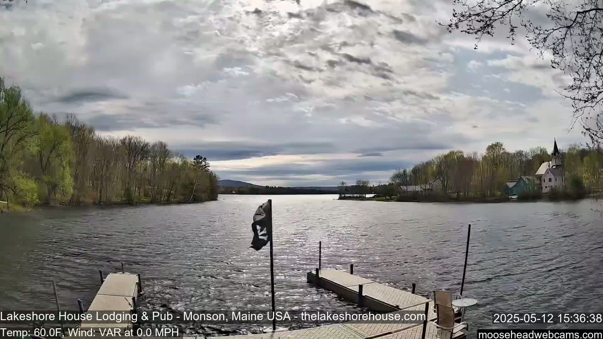 A calm lake with two docks is seen under a partly cloudy sky, with trees and buildings visible on the opposite shore.
