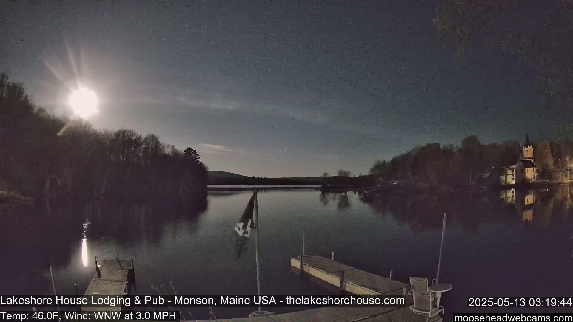 A bright moon illuminates a calm lake at night, reflecting the lights of buildings on the shore and a flagpole on a dock.