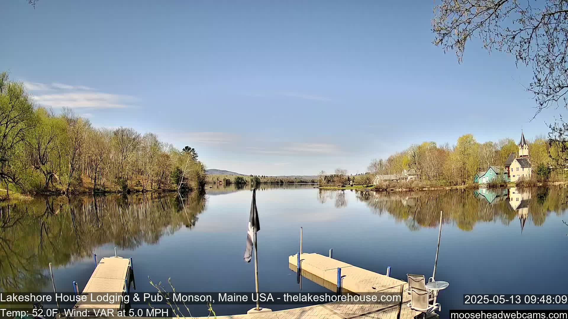 Calm, clear water reflects a tranquil lakeside scene of trees budding in spring under a blue sky, with two docks in the foreground.