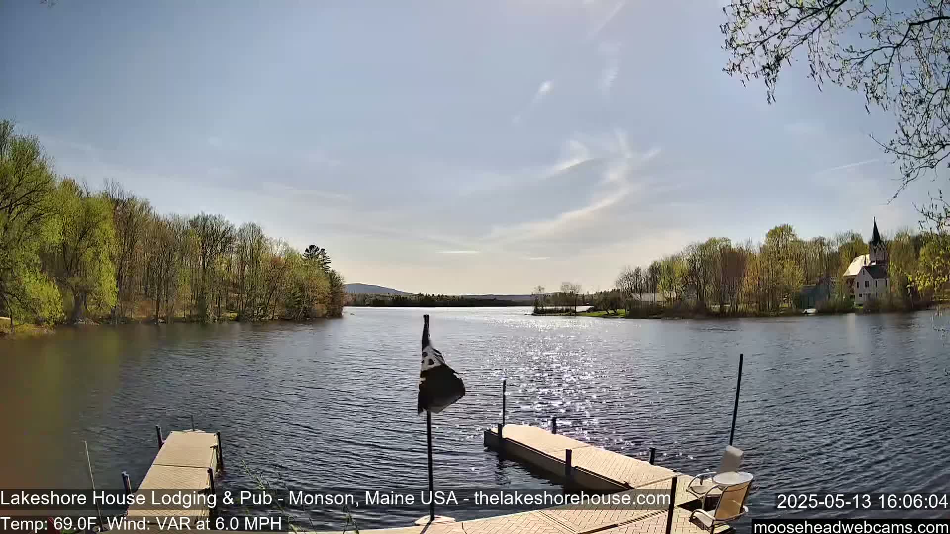 A calm lake with a dock and a flagpole, under a mostly sunny sky, is surrounded by trees and a building on the far shore.