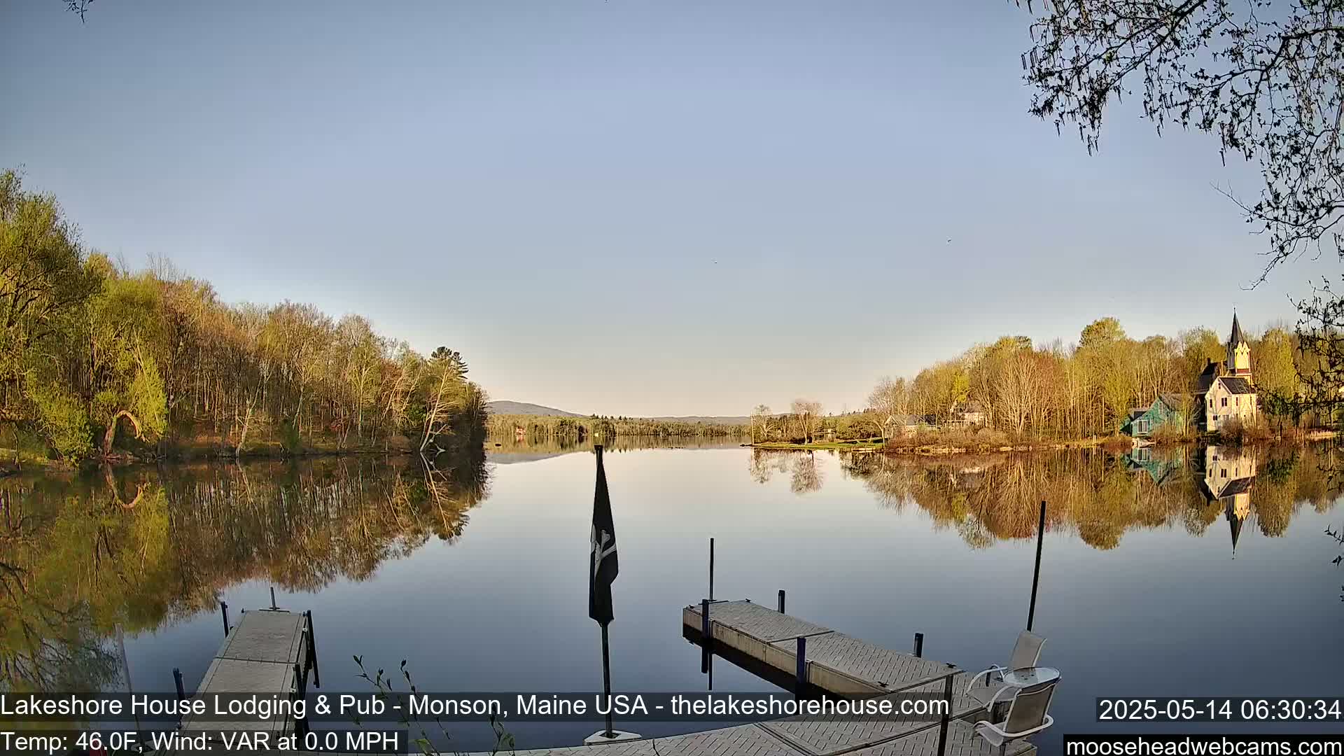 A calm lake reflects the trees and buildings on its shores under a clear, light blue sky.
