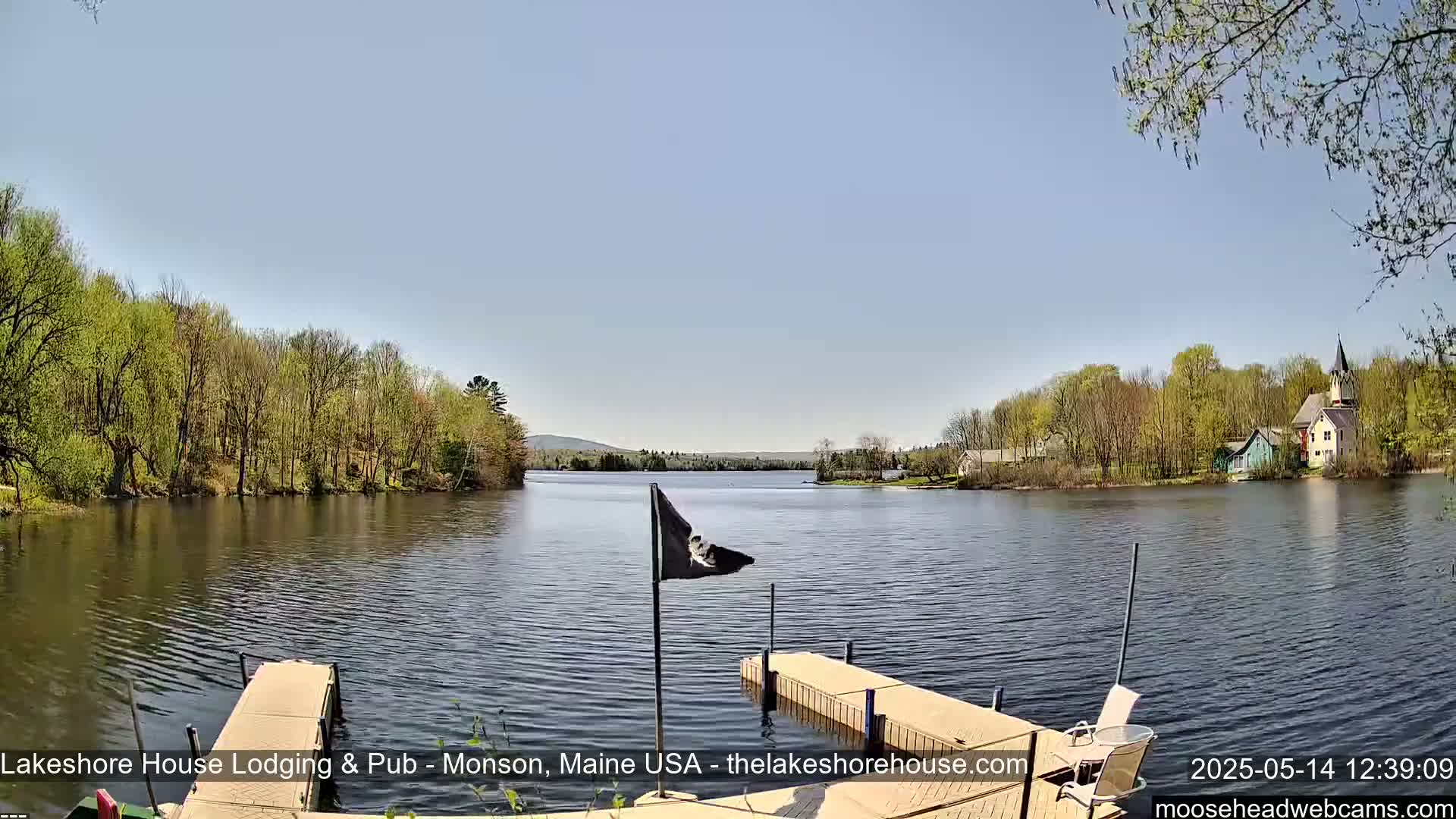 A calm lake is viewed from a dock on a sunny day, with lush green trees lining the shoreline and buildings visible in the distance.