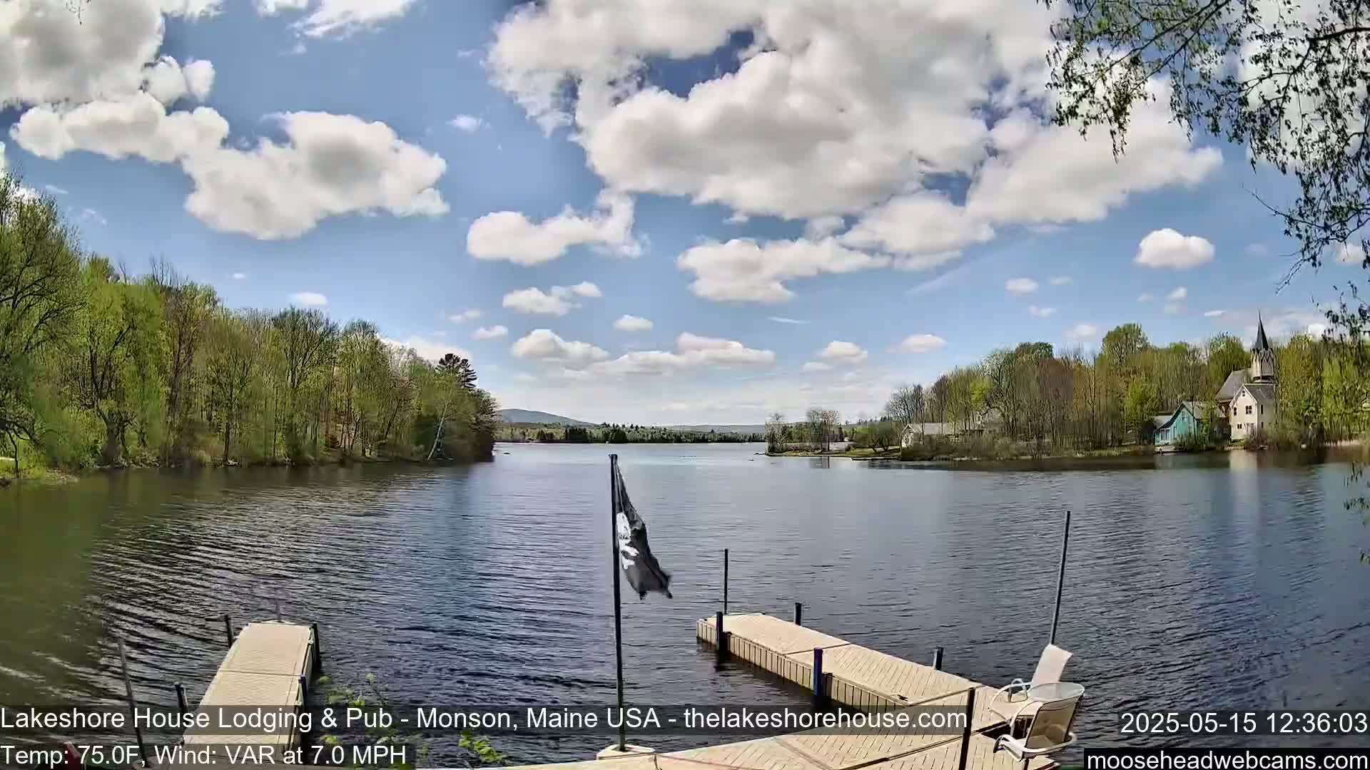 A calm lake is seen from a dock on a partly cloudy day, with lush green trees lining the shore and a few buildings visible in the distance.