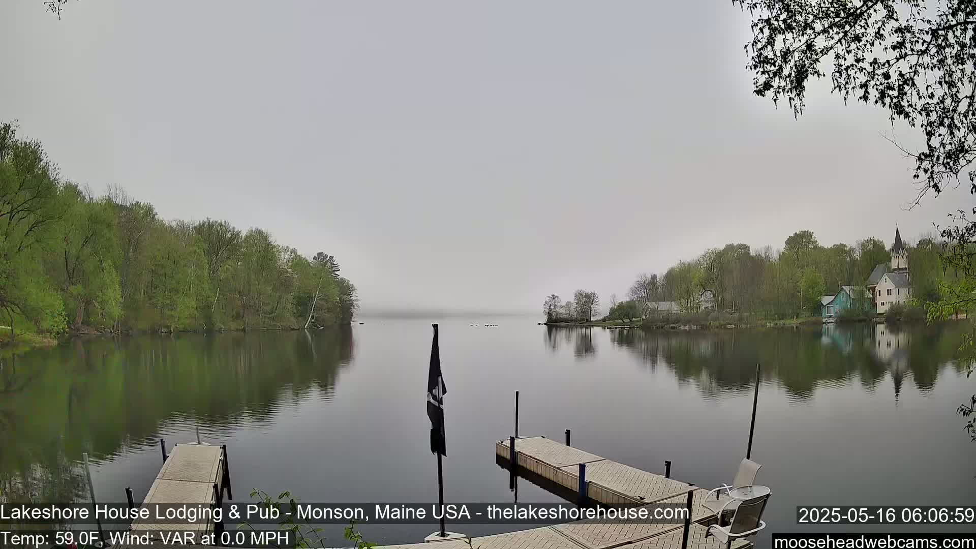 A calm, foggy lake is seen from a dock, with green trees lining the shores and buildings visible in the distance.