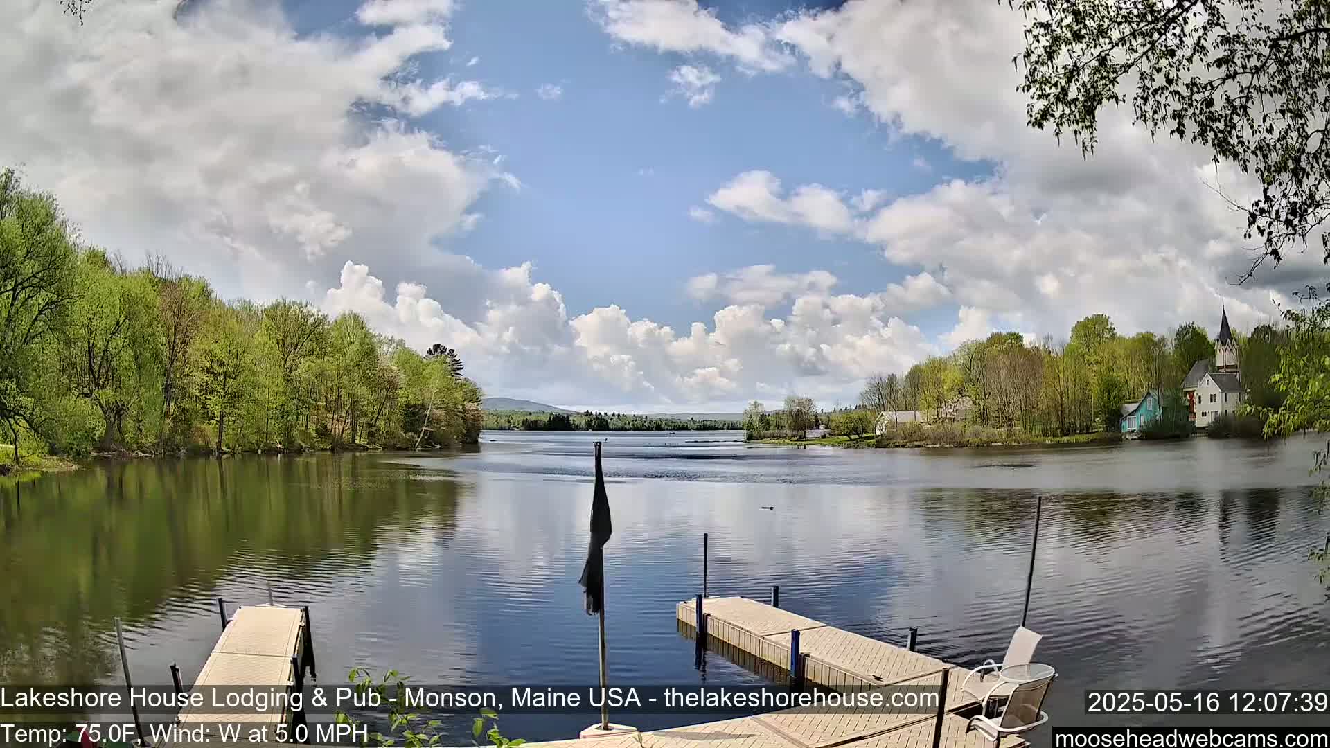A calm lake with a wooden dock and a flagpole is surrounded by lush green trees and buildings under a partly cloudy sky.