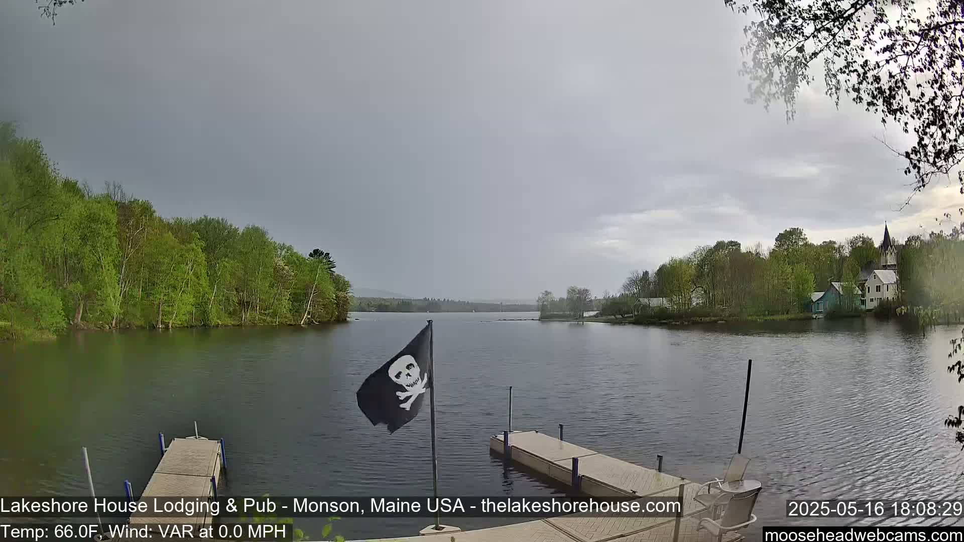 A calm lake under an overcast sky is viewed from a dock, with green trees lining the shore and a pirate flag on a post in the foreground.