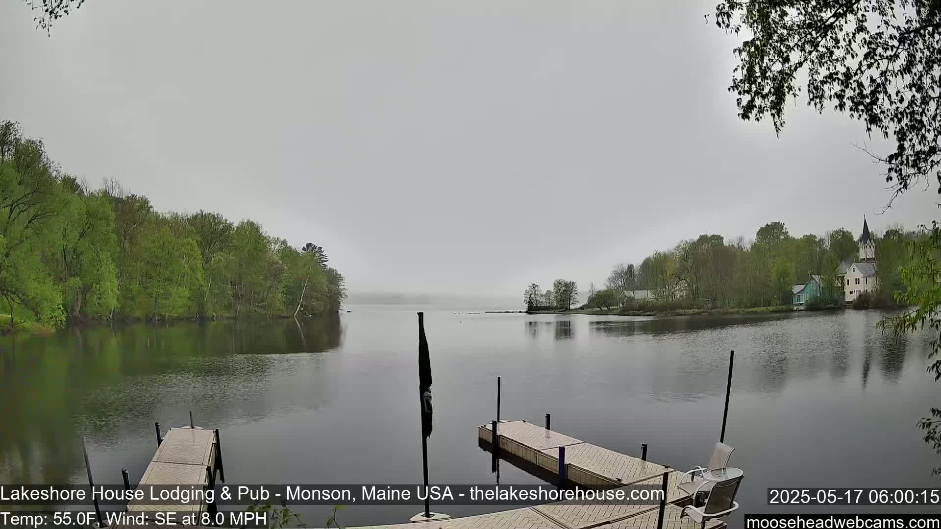 A calm lake is seen from a dock on an overcast day, with trees and buildings lining the far shore.