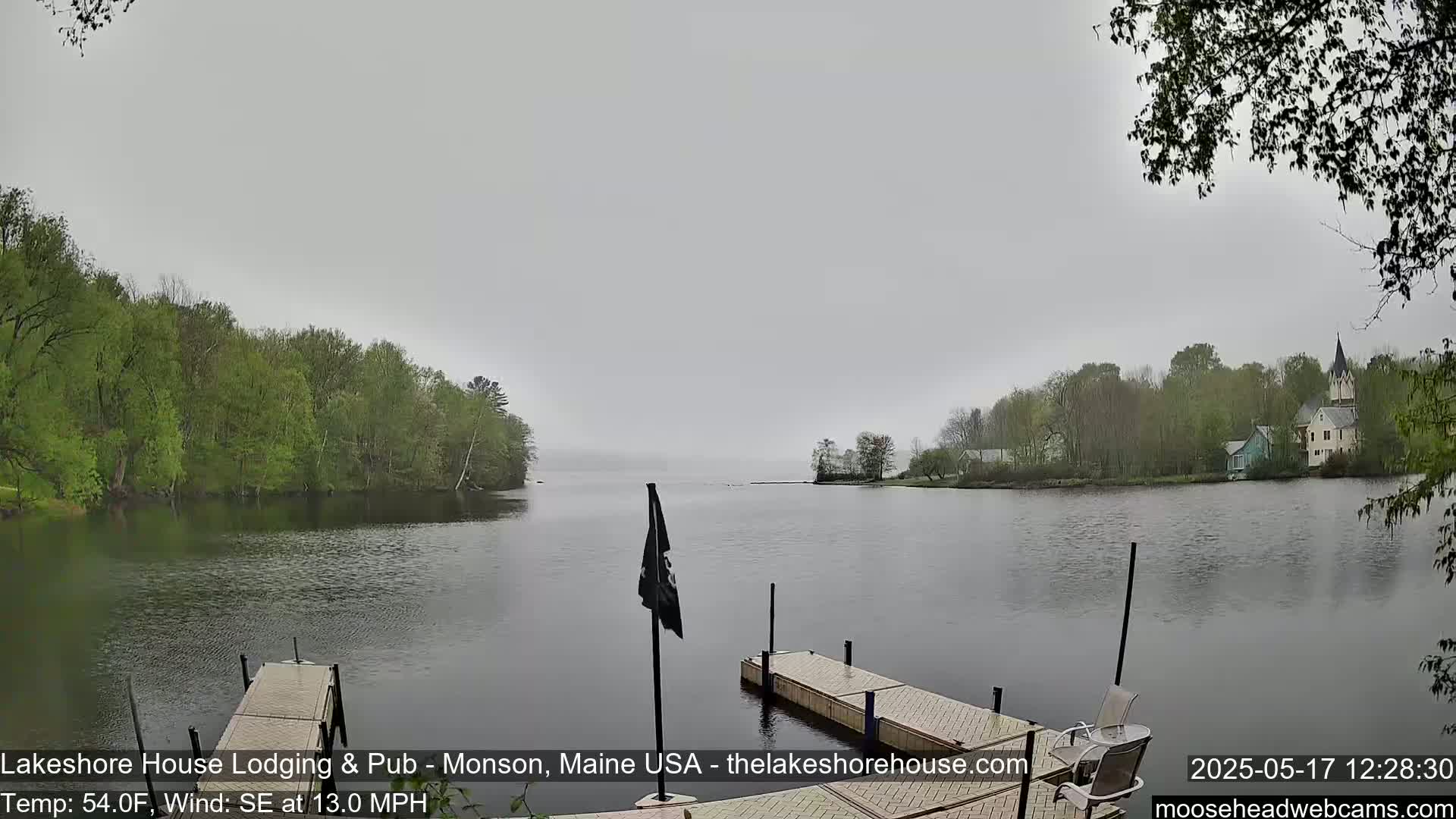 A calm lake under an overcast sky is viewed from a dock, with green trees lining the shore and buildings visible in the distance.