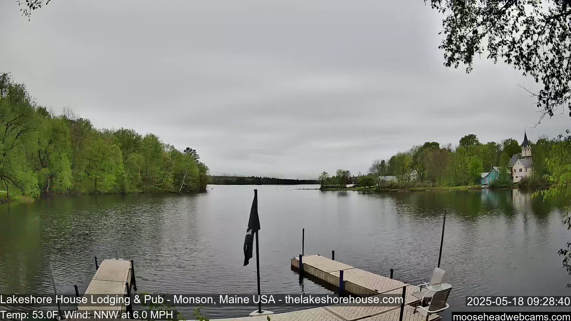 A calm lake under an overcast sky is viewed from a dock, with green trees lining the shore and buildings visible in the distance.
