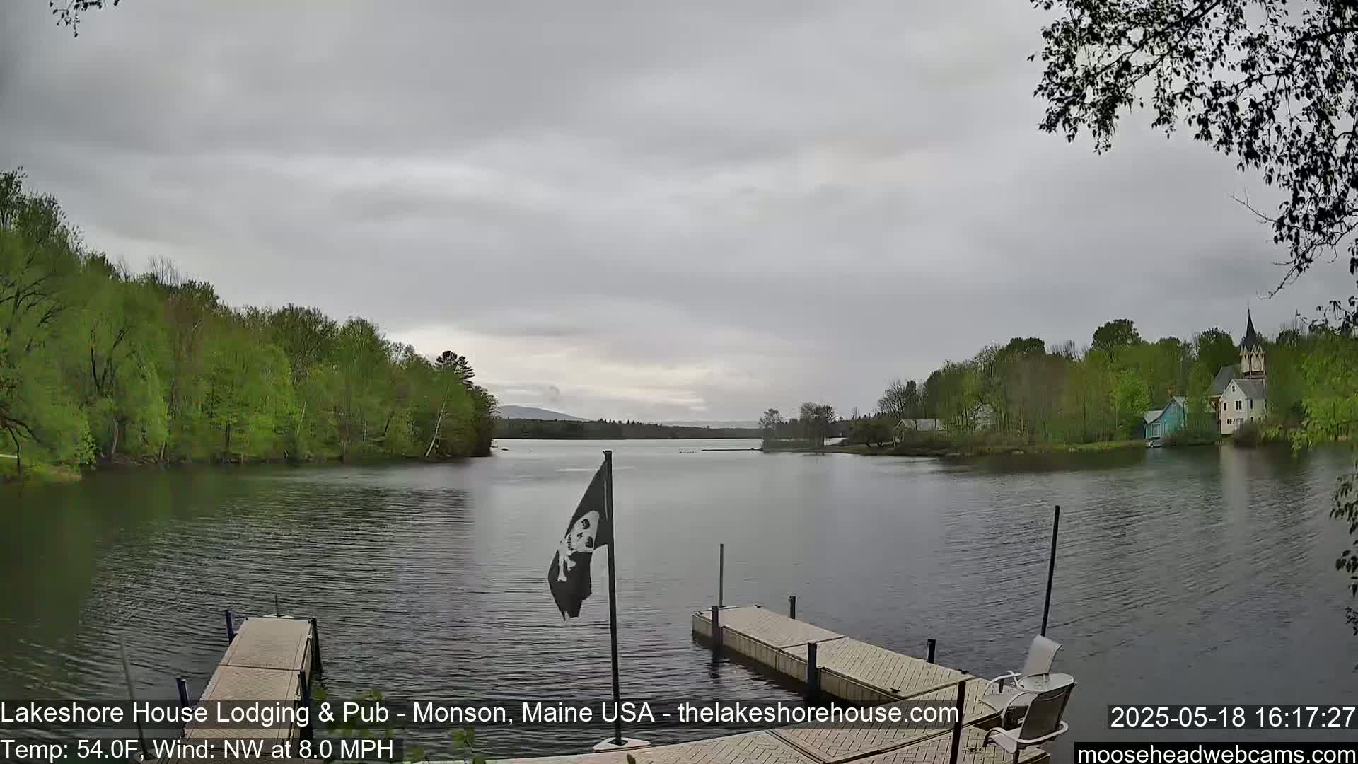 A calm lake under an overcast sky is seen from a dock, with green trees lining the shoreline and a few buildings visible in the distance.