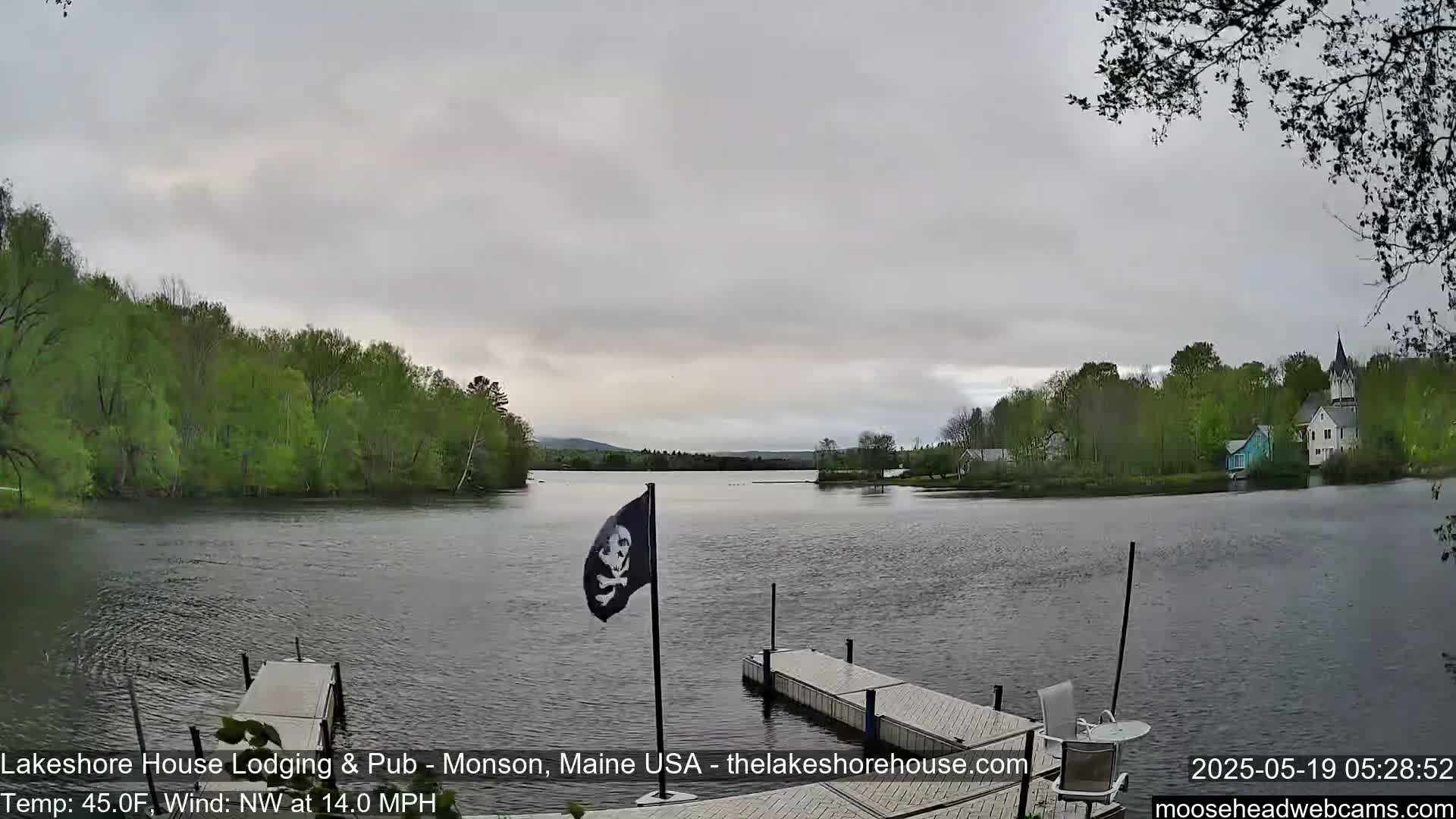 A calm lake with green trees lining its shores, under an overcast sky, with a pirate flag flying from a dock.