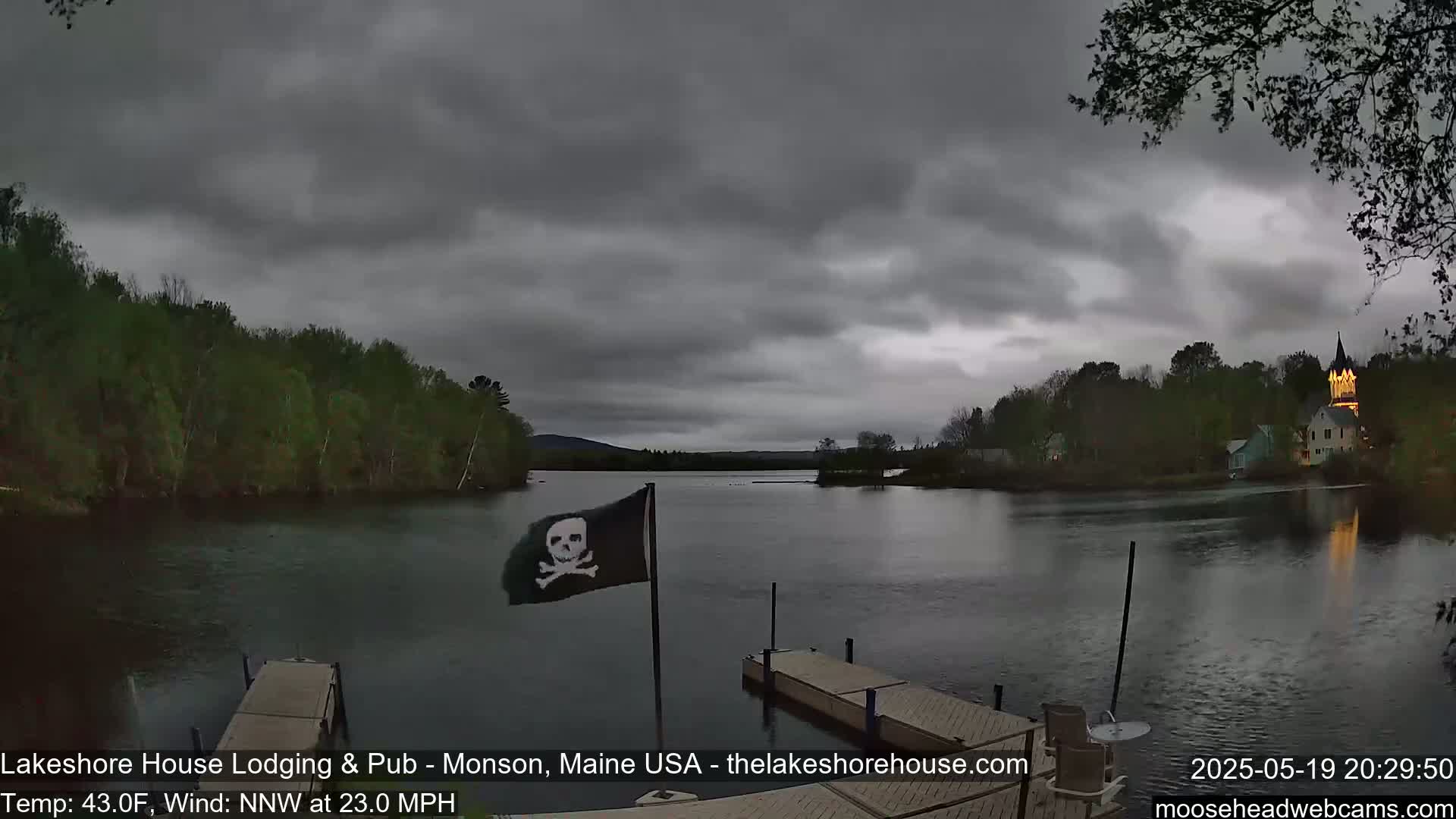 A dark gray, overcast sky hangs over a calm lake with a small dock, trees lining the shore, and a building with lights visible in the distance; a pirate flag flies from a pole near the dock.
