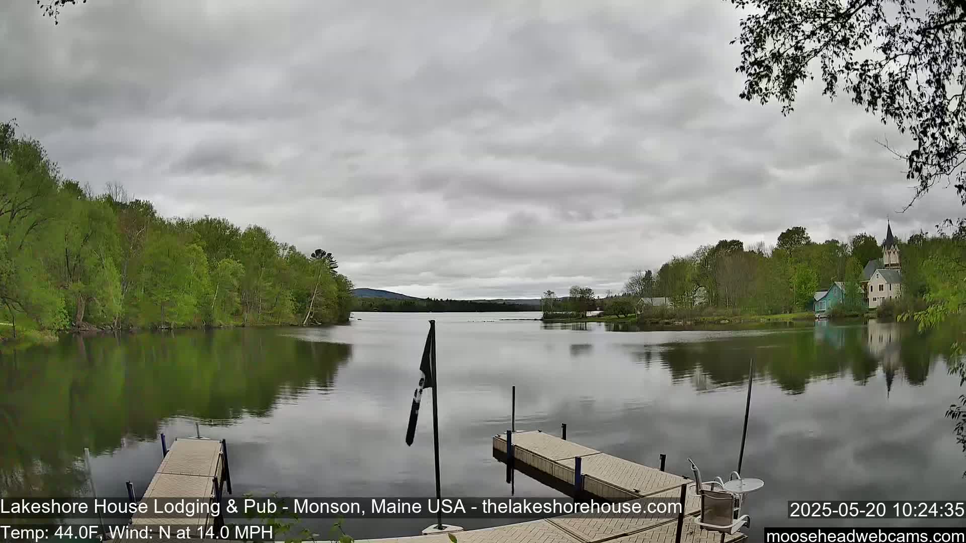 A calm lake with a wooden dock in the foreground, surrounded by green trees and buildings under a cloudy sky.