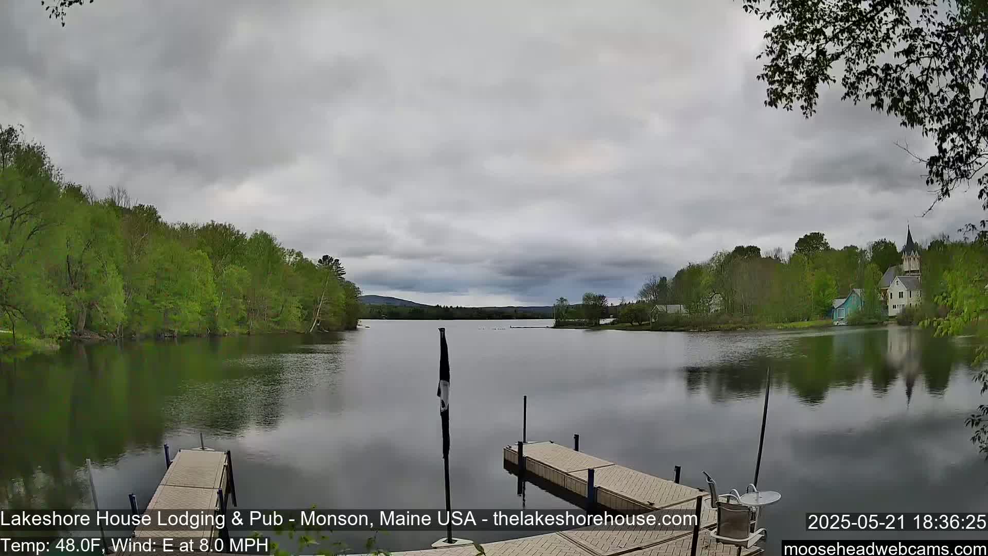 A calm lake under an overcast sky is viewed from a dock, with lush green trees lining the shore and buildings visible in the distance.