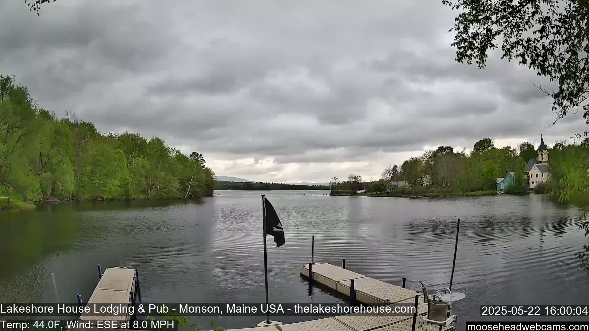 A calm lake, under a cloudy sky, is viewed from a dock with a small flagpole, showing green trees and buildings along the shoreline.