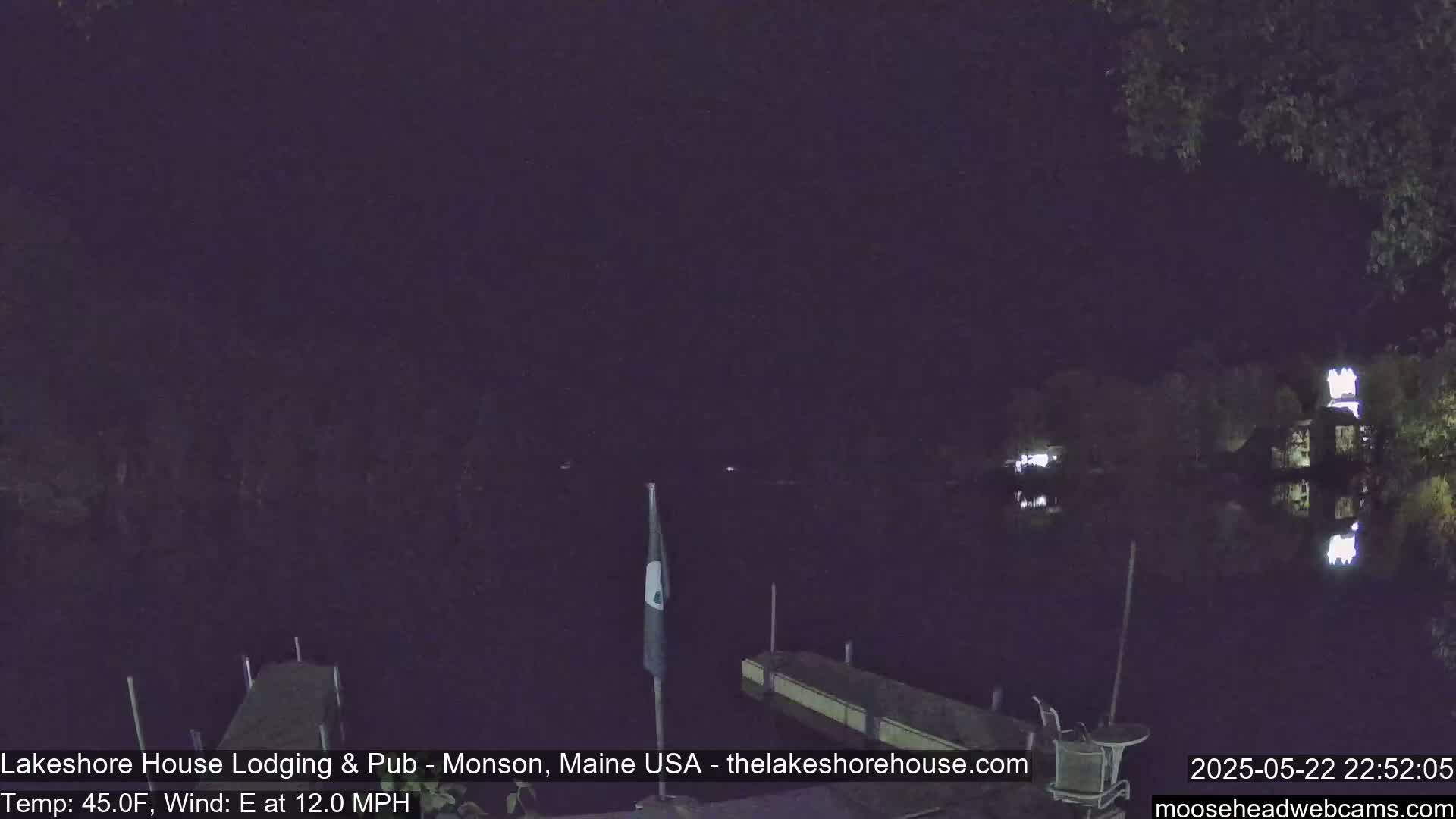 A nighttime view of a calm lake, with a dock and flagpole in the foreground, under a dark sky with an east wind.