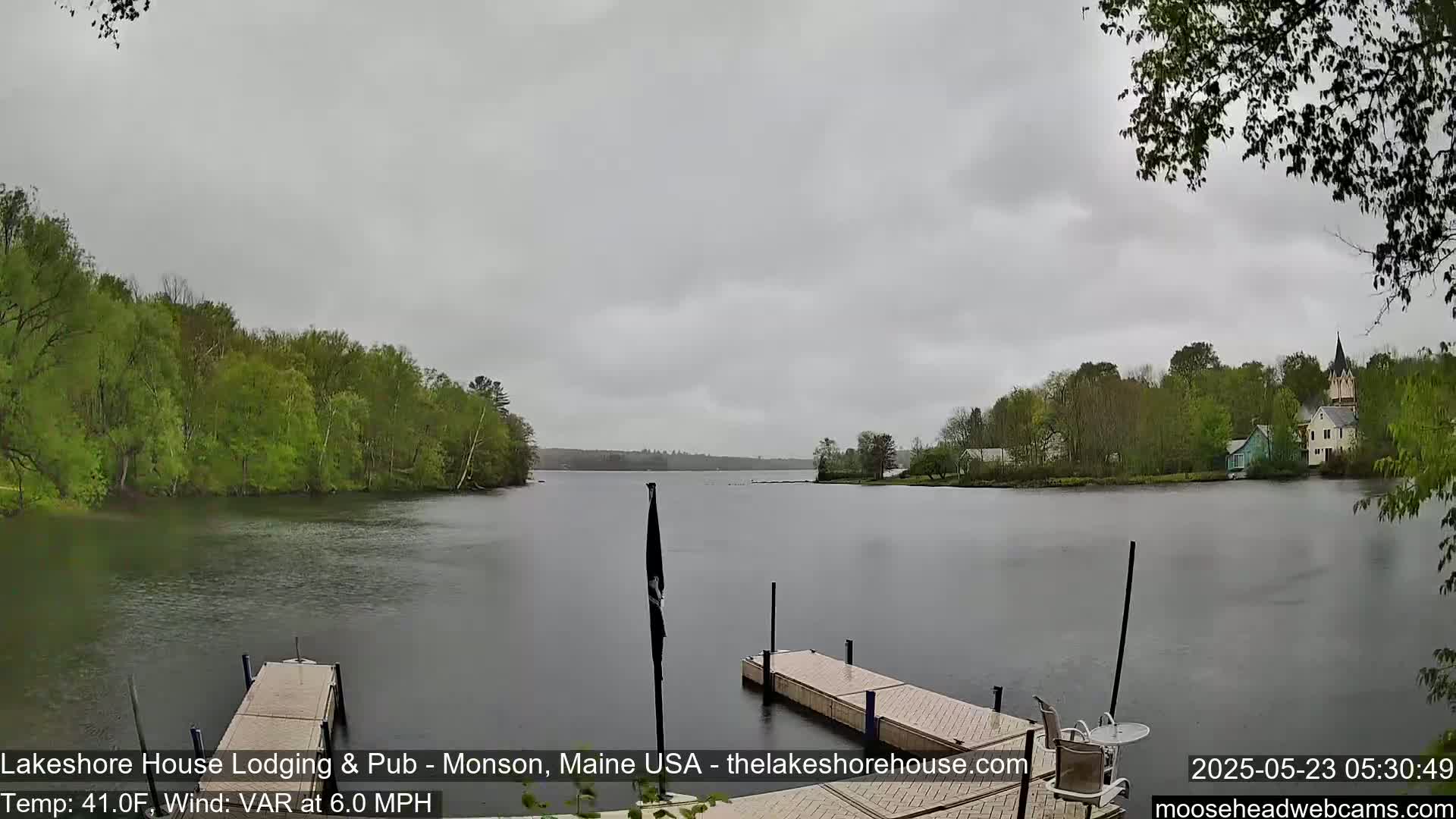 A calm lake under an overcast sky is viewed from a dock, with green trees lining the shore and buildings visible in the distance.