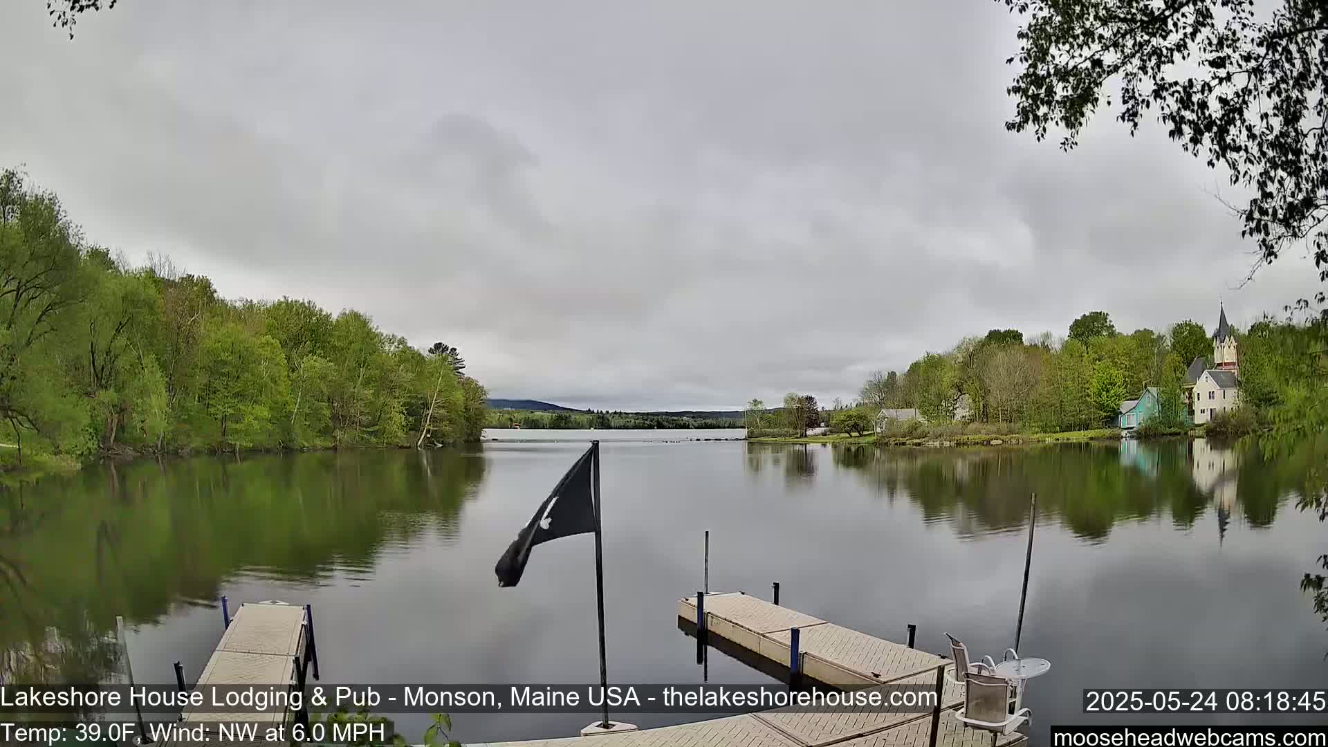 A calm lake with a wooden dock, surrounded by lush green trees and buildings under an overcast sky.