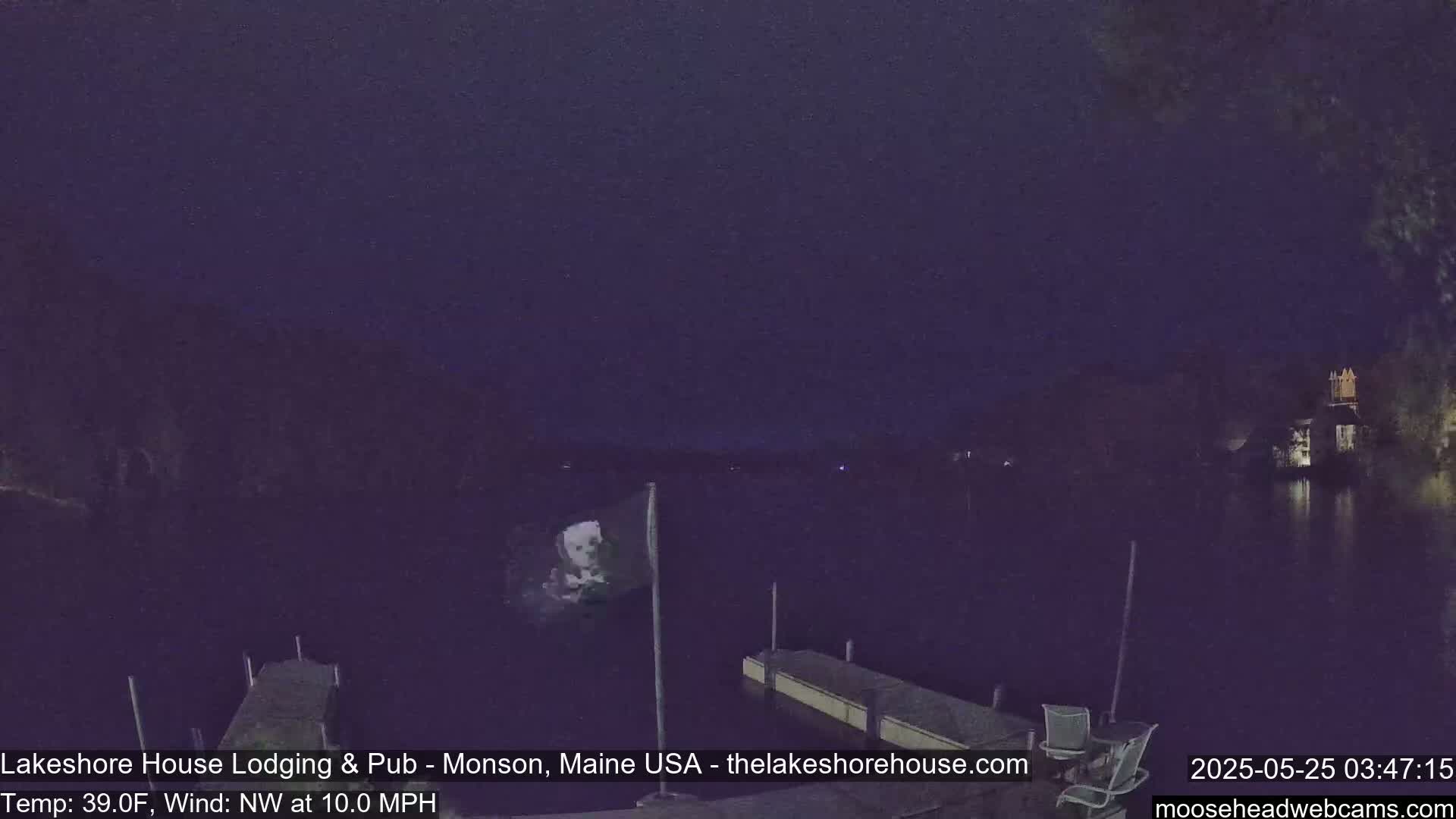 A nighttime view of a dark lake with two docks, a building in the distance, and a flagpole with a flag in the water under a dark, mostly clear sky, with a northwest wind at 10 mph and a temperature of 39°F.