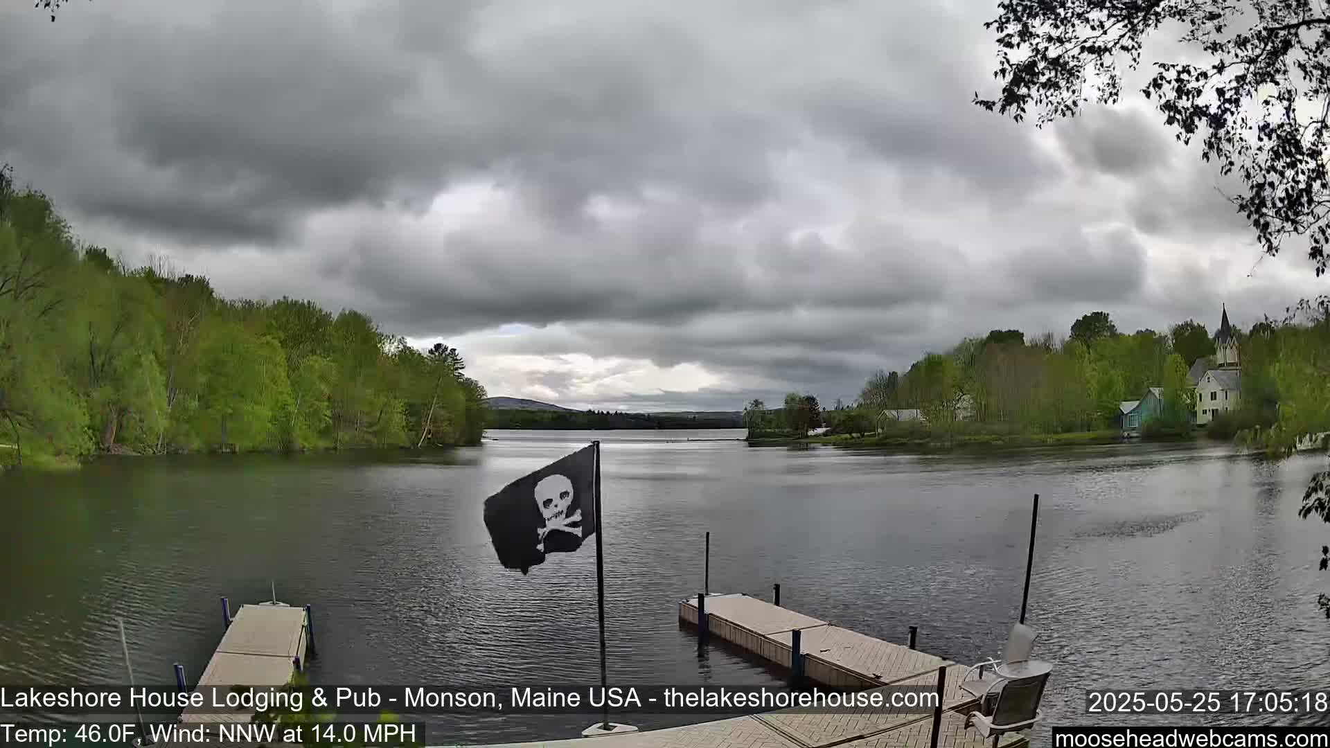 A calm lake under a cloudy sky is seen from a dock, with a small pirate flag on a pole and green trees lining the shoreline.