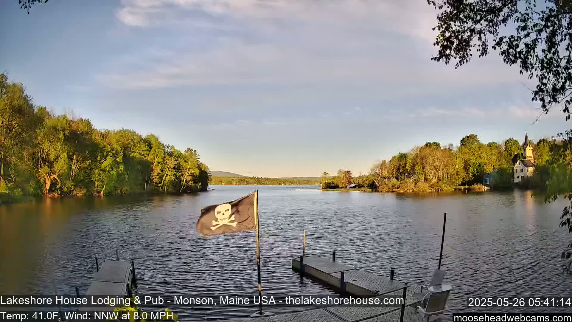A calm lake with a small dock and a pirate flag in the foreground, surrounded by lush green trees and a building on the far shore under a partly cloudy sky.