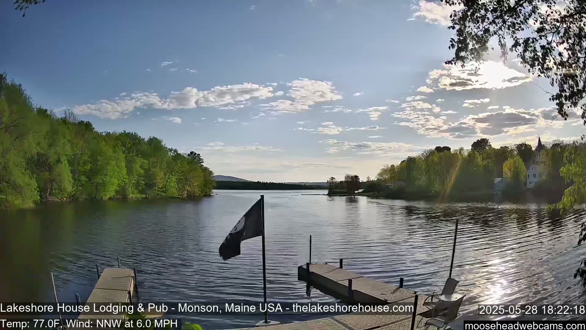 A calm lake with a wooden dock and green trees lining the shore under a partly cloudy sky.